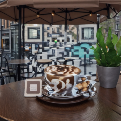 Steaming cup of coffee with artful latte design, placed on a café table with a blurred city street scene in the background.